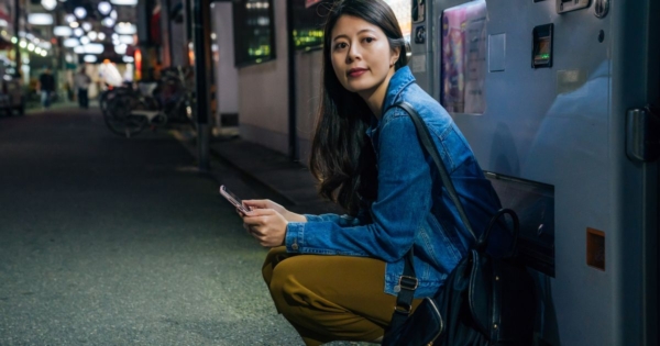 young Asian female student crouching and looking at phone while waiting for a ride at night